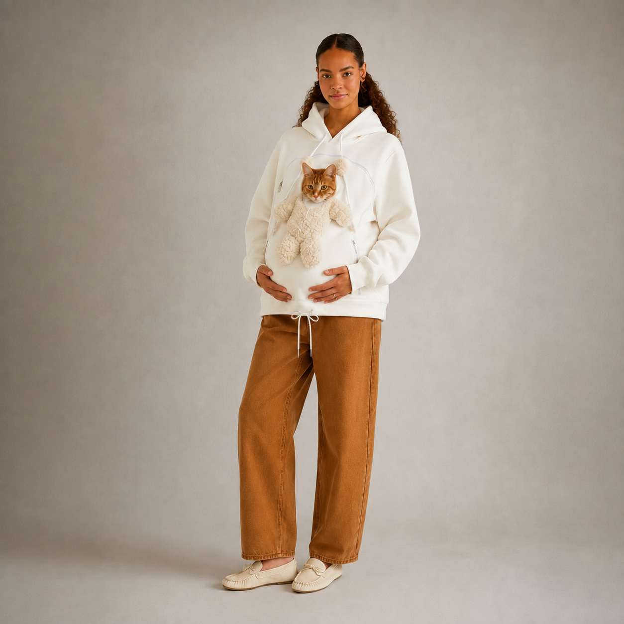 A woman stands against a plain light gray studio background wearing a white hoodie with a cat stuffed inside the front pocket, paired with wide-leg brown pants and cream slip-on shoes.