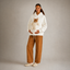 A woman stands against a plain light gray studio background wearing a white hoodie with a cat stuffed inside the front pocket, paired with wide-leg brown pants and cream slip-on shoes.