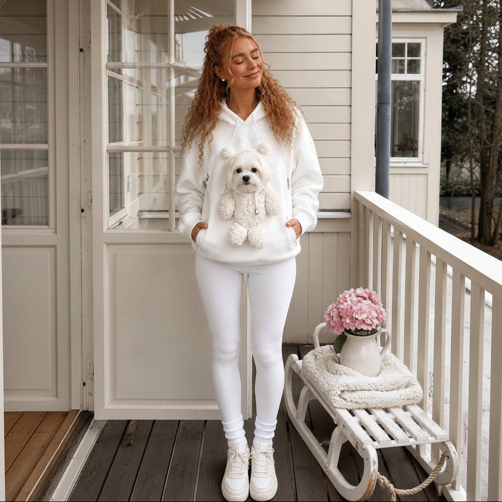 A woman stands on a porch wearing a white hoodie with a plush white dog teddy no the pocket, paired with white leggings and white sneakers. A small white dog is also sitting on the pocket next to a bench with pink flowers.