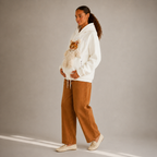 A woman stands in a studio wearing a white hoodie with a plush white bear on the front cat pocket, paired with wide-leg brown pants and cream sneakers. The background is a solid light gray with soft diagonal shadows.