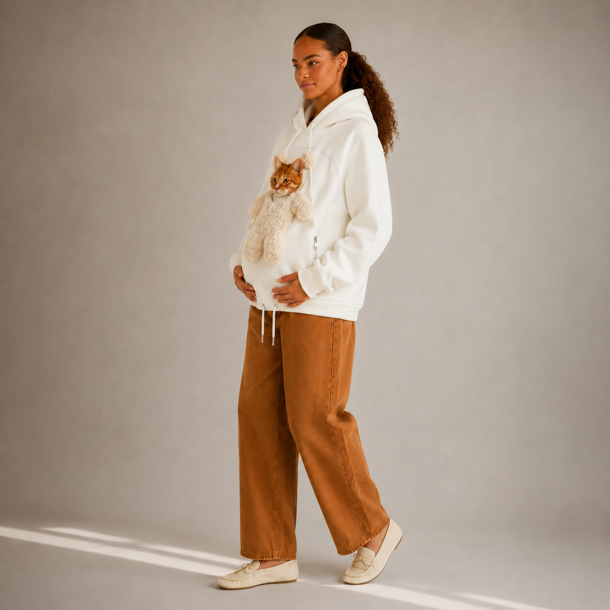 A woman stands in a studio wearing a white hoodie with a plush white bear on the front cat pocket, paired with wide-leg brown pants and cream sneakers. The background is a solid light gray with soft diagonal shadows.