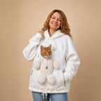 A woman wearing a white fleece hoodie with a plush teddy bear pocket containing an orange tabby cat, posed against a solid beige studio background.