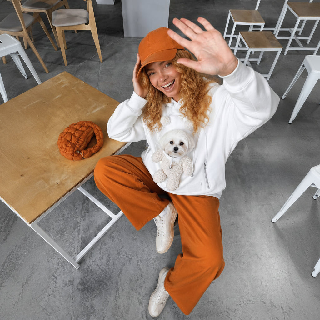 A woman wearing a white hoodie, orange pants, and an orange beanie waves while sitting on a table in a cafe, with a small white dog on her lap and an orange knit hat on the table next to her.