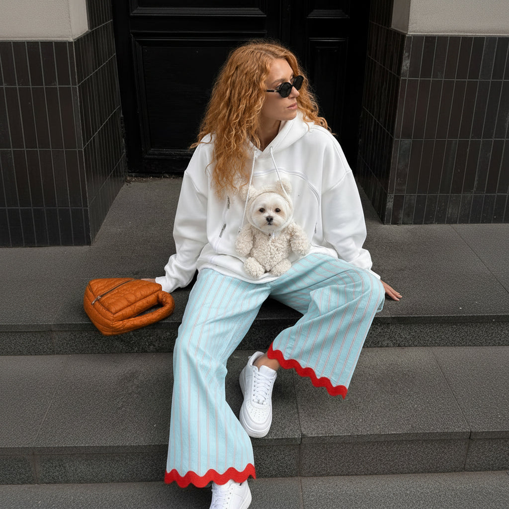 A woman with curly red hair wearing a white hoodie with a teddy bear pocket, light blue and white striped pants with a red scalloped hem, and white sneakers, sitting on stone steps with a small white dog in her pouch.