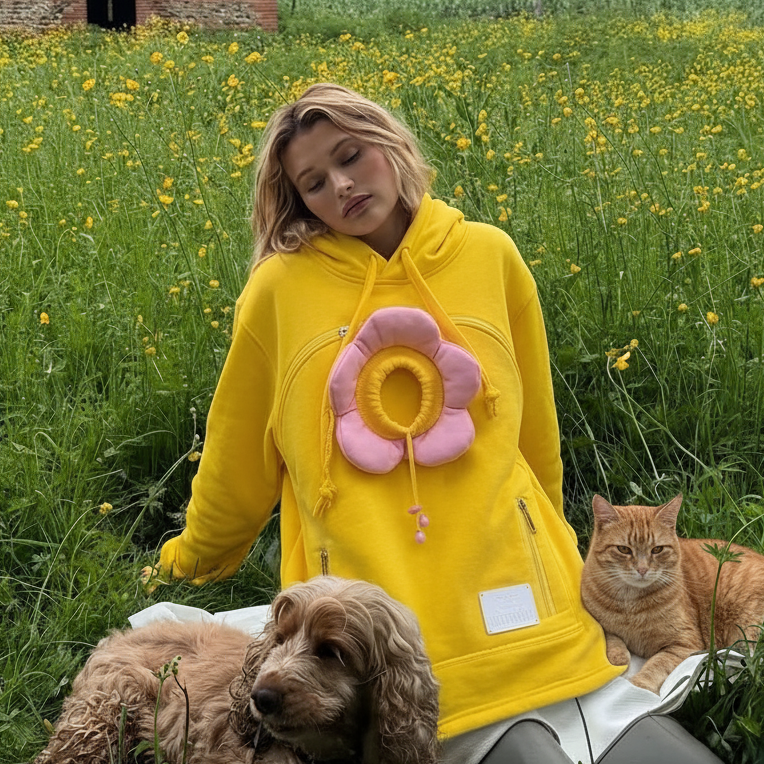 A woman wearing a bright yellow hoodie with a pink flower applique sits in a grassy field with yellow flowers, accompanied by two dogs.